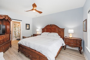 A bedroom with a wooden four-poster bed, two bedside tables with lamps, a ceiling fan, and a television on a wooden cabinet.