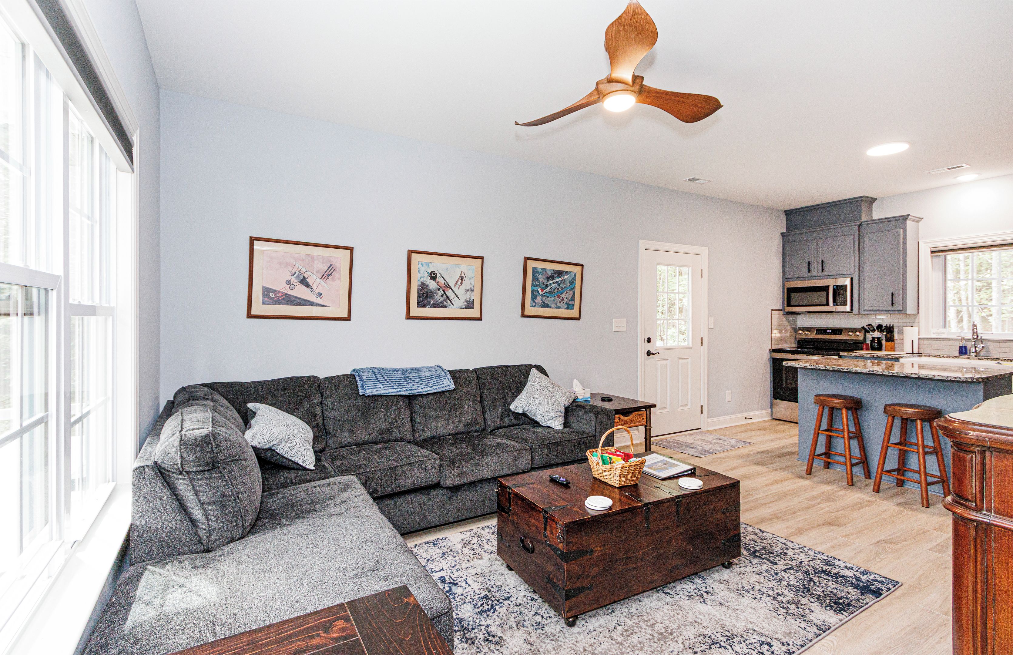 A living room with a sofa, coffee table, and a ceiling fan. There are framed pictures on the wall and a kitchen area with a microwave and bar stools.