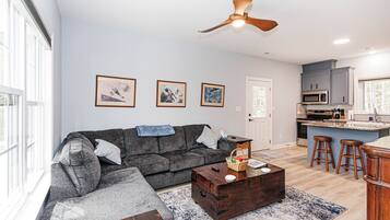A living room with a sofa, coffee table, and a ceiling fan. There are framed pictures on the wall and a kitchen area with a microwave and bar stools.