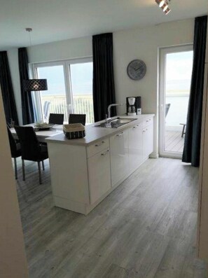 A modern kitchen with a large island, a dining area with chairs, and a view of the sea through the windows. - 47.6.n Flashing Light (Wangerland)