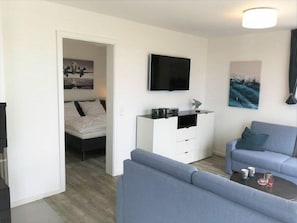 A modern living room with a sofa, a coffee table, a flat-screen TV mounted on the wall, and a bed in the adjacent room. - 47.6.n Flashing Light (Wangerland)