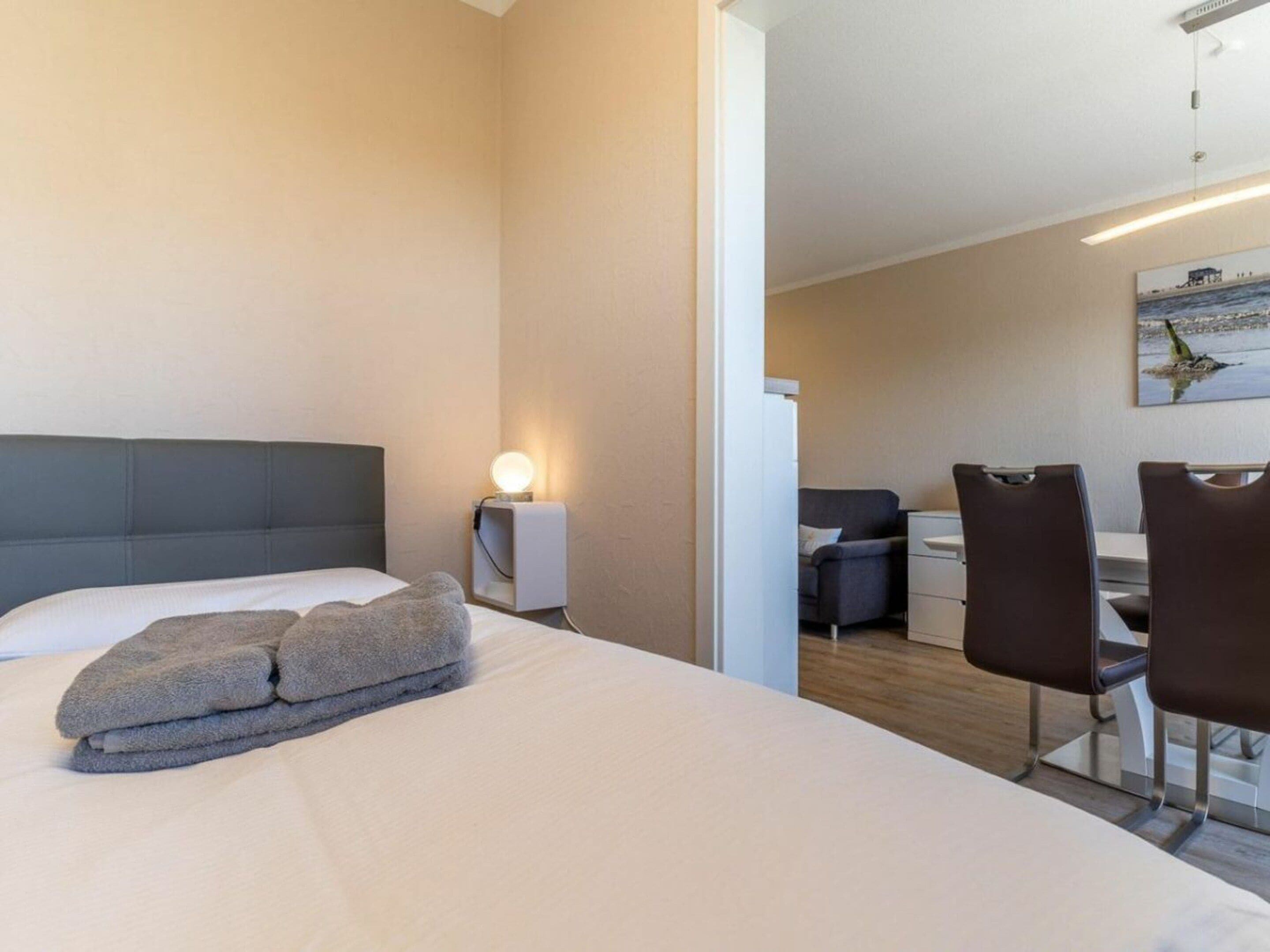 A hotel room with a bed, a bedside lamp, a desk with chairs, and a painting on the wall.