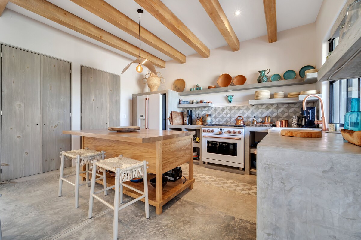Een moderne keuken met houten balken, een centraal eiland, barstoelen en diverse keukengerei op planken.