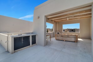 An outdoor patio area with a built-in kitchen, a wooden bench, and a view of neighboring buildings. - Baja Sage (El Pescadero)