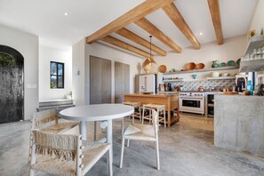 A modern kitchen with wooden beams, a round dining table, wicker chairs, and a built-in oven.