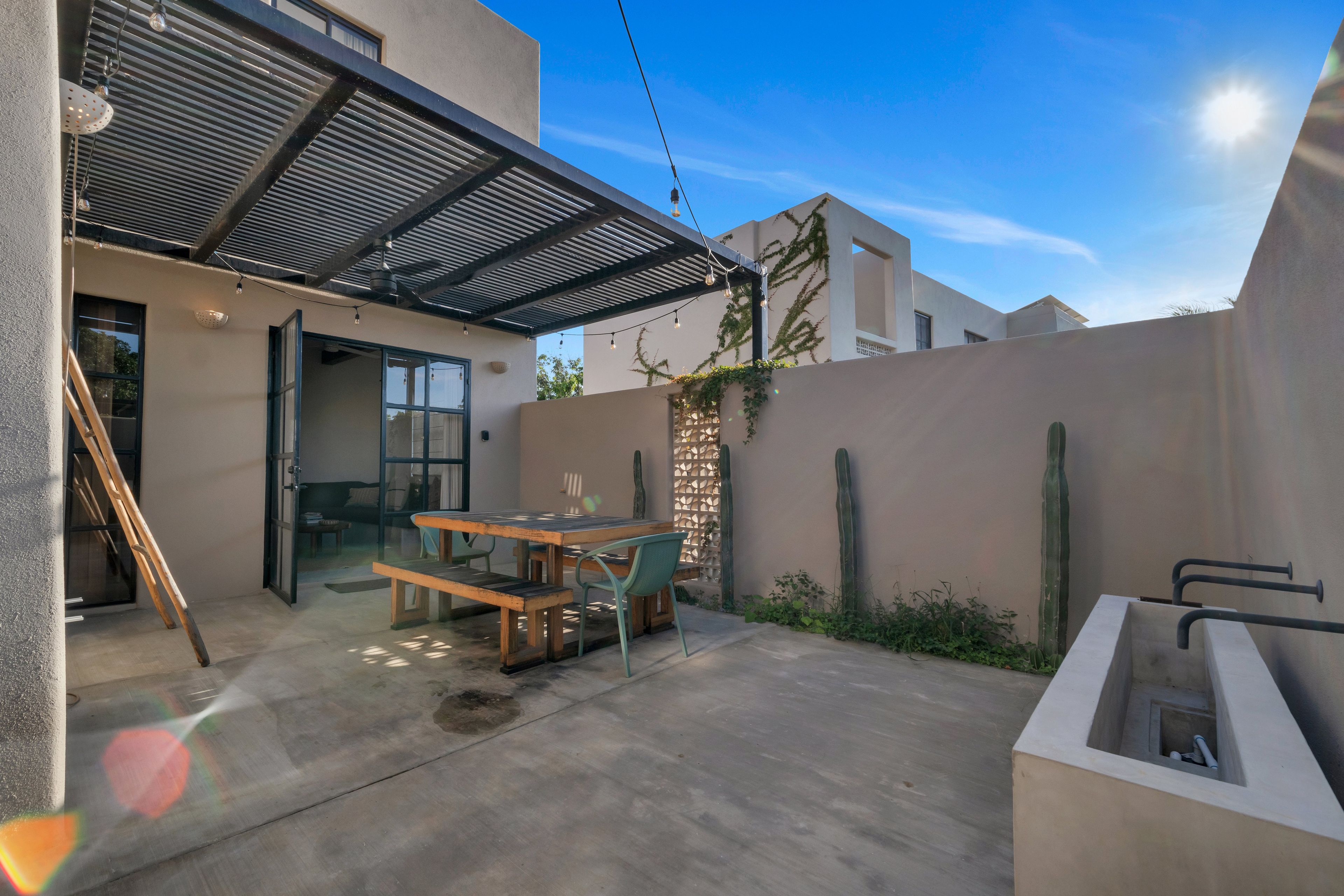 An outdoor patio with a table and chairs, a pergola with string lights, a ladder, and a wall with climbing plants.