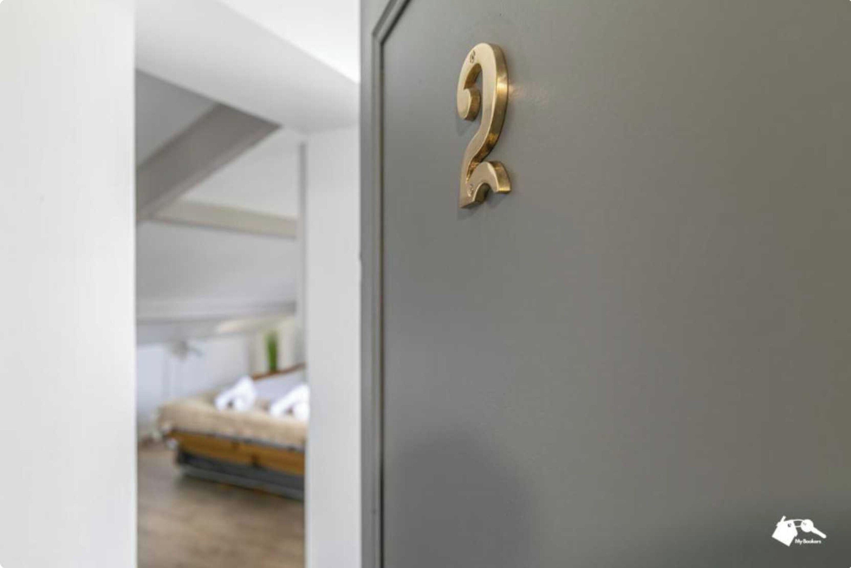 A room with a grey door marked with the number '2' and a glimpse of a bed in the background.