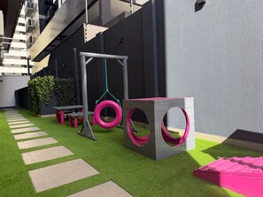 A modern outdoor play area with artificial grass, a swing set, a tire swing, and a playhouse. - AL - BXSU - Box Studio  (Maceió)