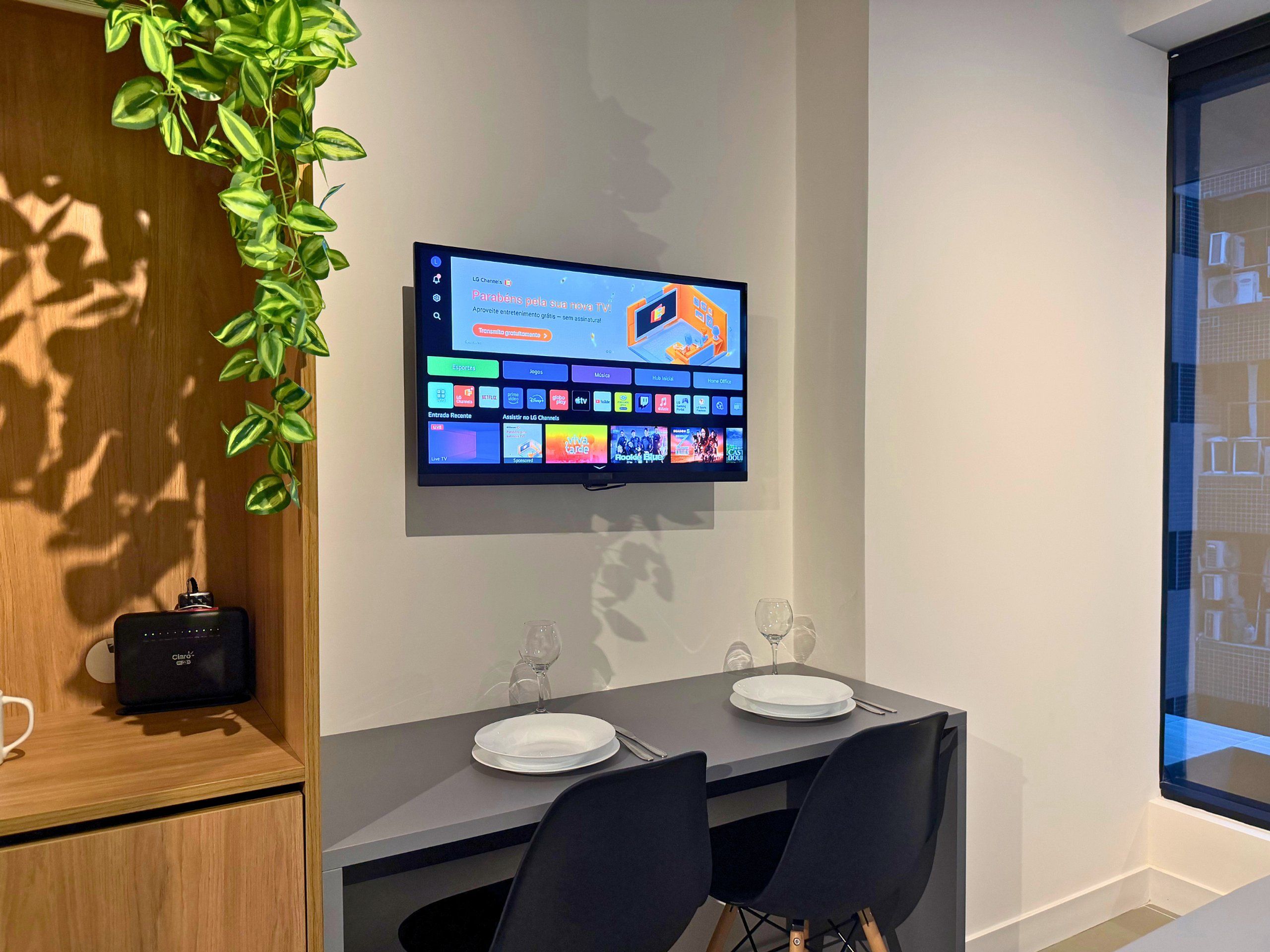 Uma sala de jantar moderna com uma televisão de tela plana fixada na parede exibindo um guia de programação. Há uma mesa de jantar com cadeiras, uma planta e um pequeno alto-falante em um armário de madeira.