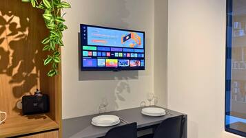 Uma sala de jantar moderna com uma televisão de tela plana fixada na parede exibindo um guia de programação. Há uma mesa de jantar com cadeiras, uma planta e um pequeno alto-falante em um armário de madeira.