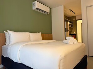 Habitación de hotel con cama, ropa de cama blanca, unidad de aire acondicionado y baño visible a través de una puerta abierta.