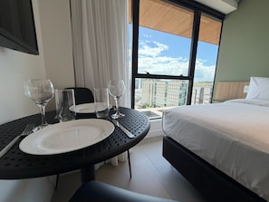 Habitación de hotel con cama, una mesa de comedor con platos y vasos, y vistas de la ciudad a través de amplios ventanales.