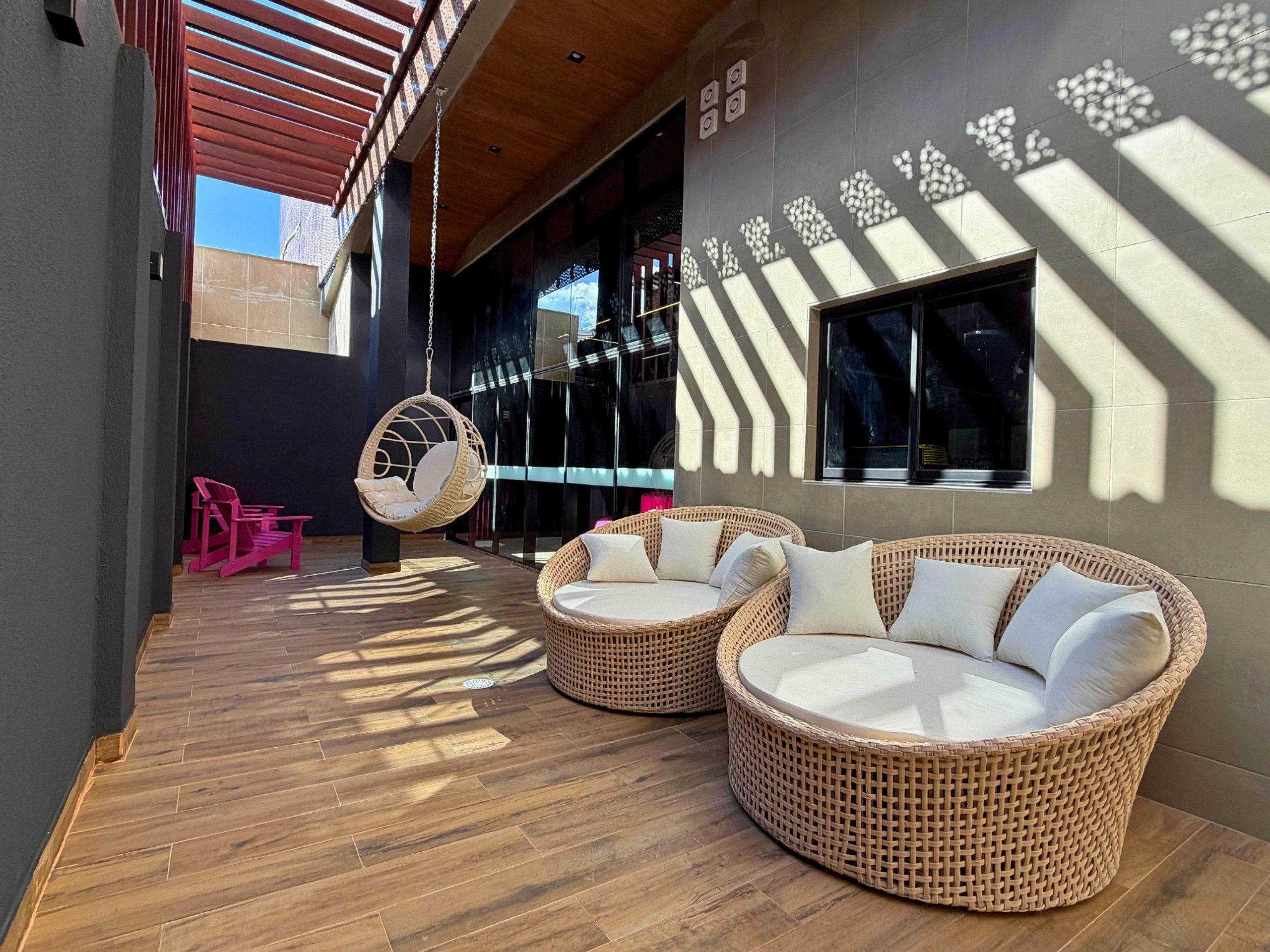 An outdoor seating area with wicker furniture and a hanging chair, a covered walkway, and a building with patterned wall design.