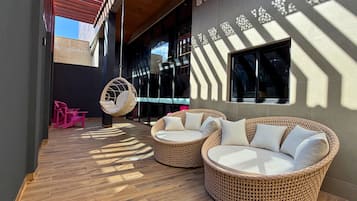 An outdoor seating area with wicker furniture and a hanging chair, a covered walkway, and a building with patterned wall design.
