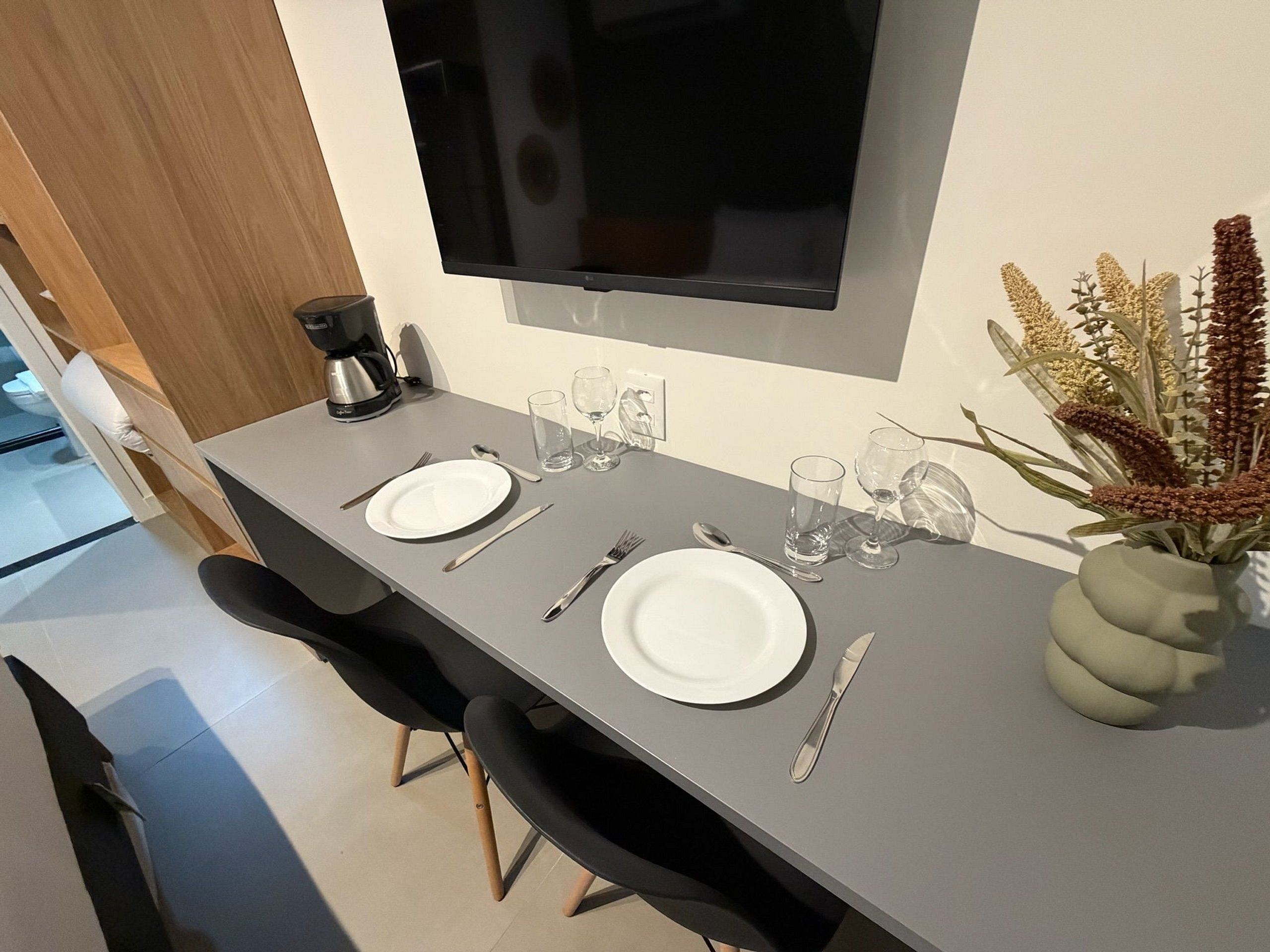 Un comedor con una mesa preparada para dos, un televisor de pantalla plana montado arriba y un florero con flores secas a la derecha.