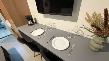 Un comedor con una mesa preparada para dos, un televisor de pantalla plana montado arriba y un florero con flores secas a la derecha.