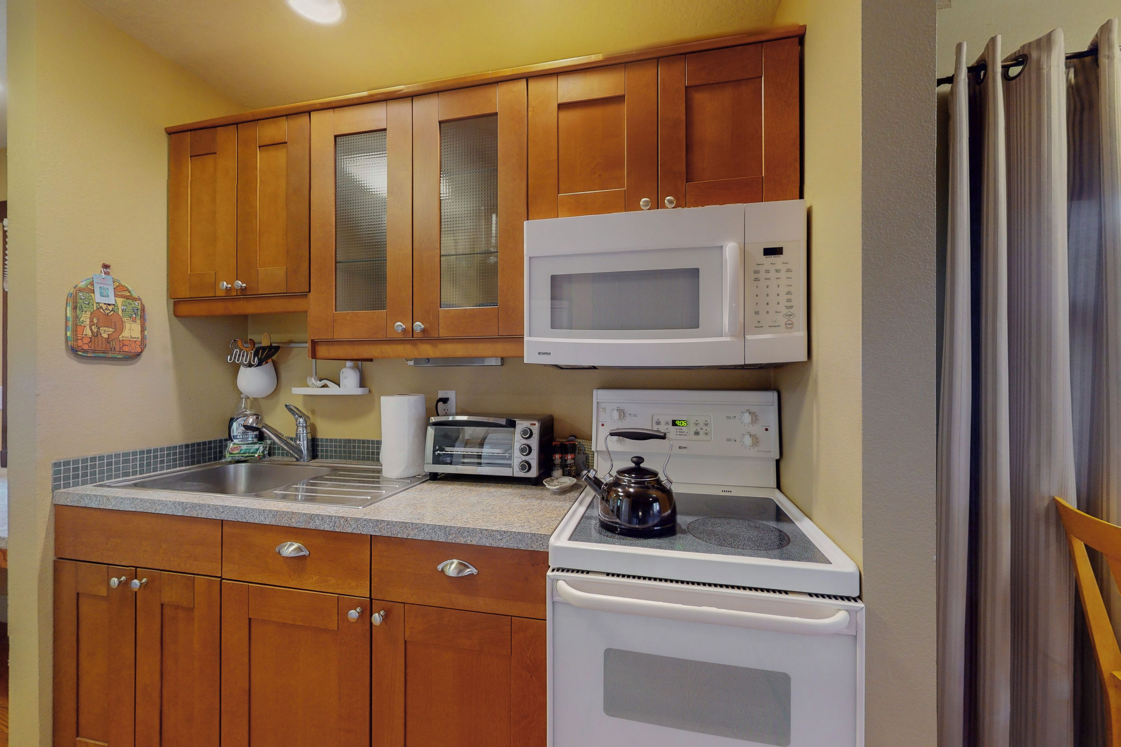 A compact kitchen with wooden cabinets, a microwave, a toaster oven, and a kettle on the stove.