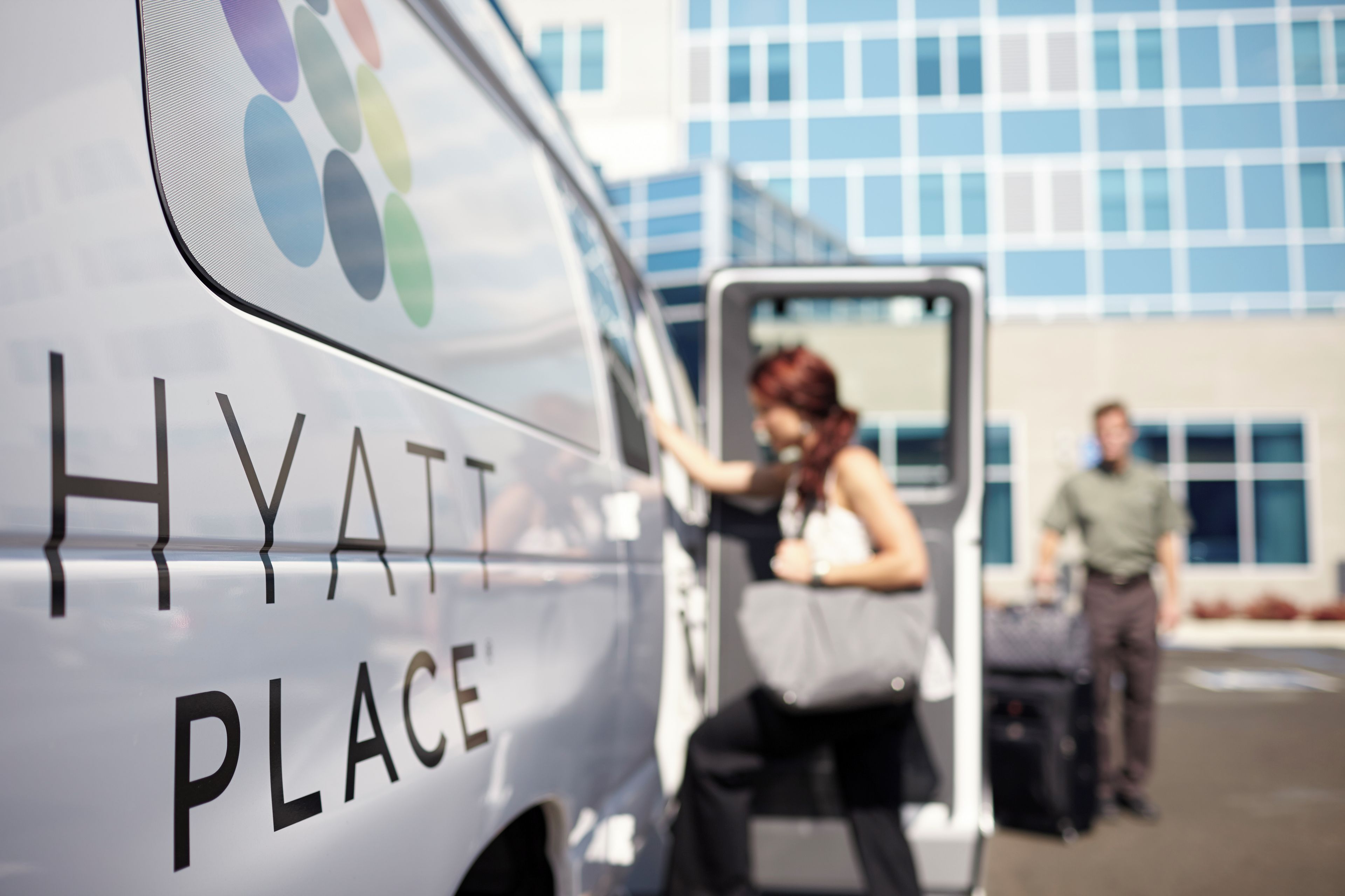 Een hotel servicebusje met 'HYATT PLACE' branding, een persoon die het busje verlaat, en een gebouw met grote ramen op de achtergrond.