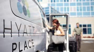 Een hotel servicebusje met 'HYATT PLACE' branding, een persoon die het busje verlaat, en een gebouw met grote ramen op de achtergrond.