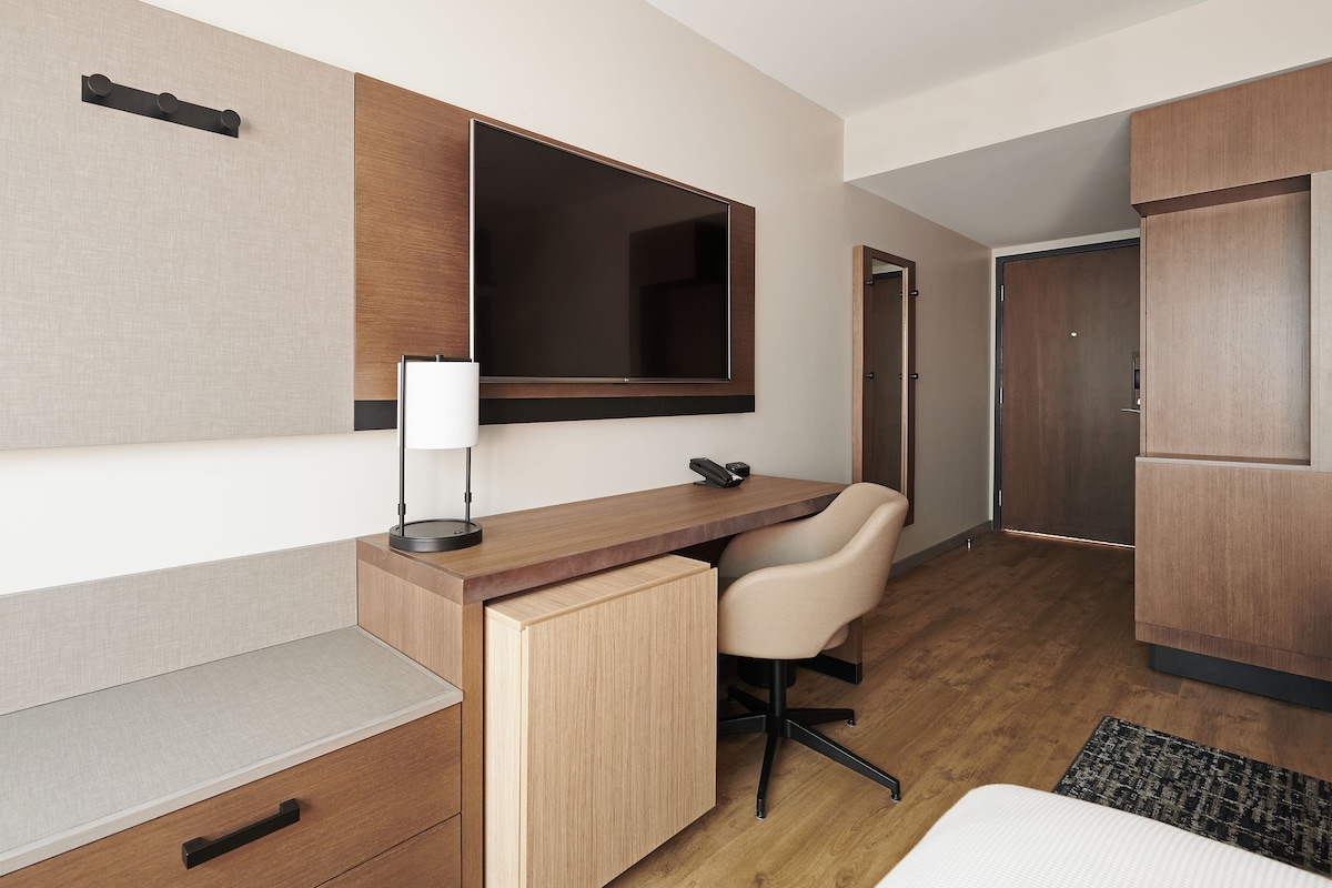 A modern hotel room with a wooden desk, a chair, a bedside table, a lamp, and a wardrobe.