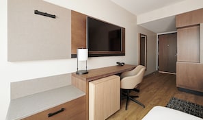 A modern hotel room with a wooden desk, a chair, a bedside table, a lamp, and a wardrobe.