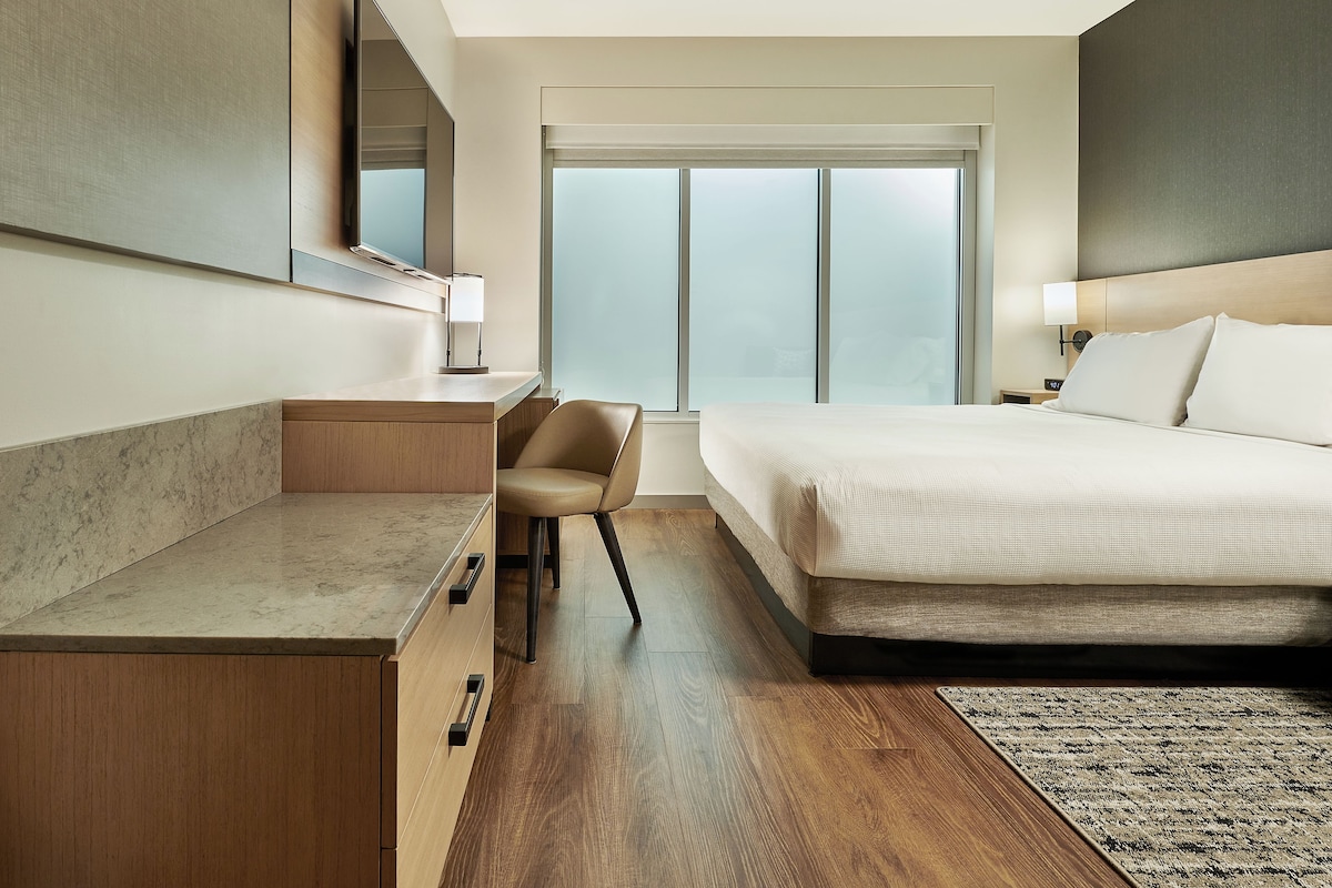 A hotel room with a bed, desk, chair, and a large window.