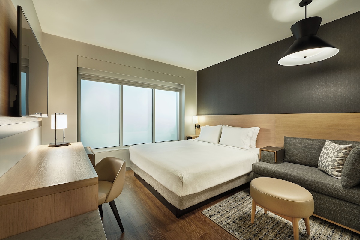 A modern hotel room with a large bed, a desk, a sofa, and a large window.