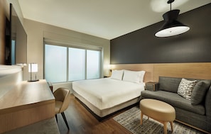 A modern hotel room with a large bed, a desk, a sofa, and a large window.