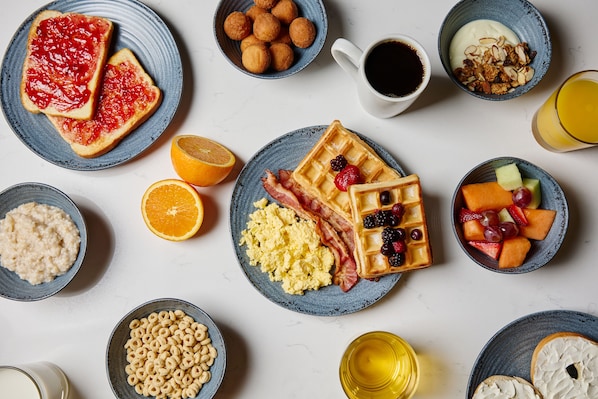 Una mesa de desayuno con tostadas, waffles, huevos revueltos, tocino y diversas frutas y cereales.