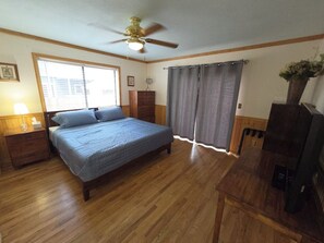 A bedroom with a bed, wooden floor, ceiling fan, window with blinds, curtains, and a television. - Serenity Lodge:5BR w/AC,5 Min/Town/Golf/Train (Williams)