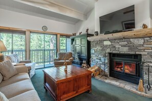 A cozy living room with a stone fireplace, comfortable seating, and a large window overlooking greenery. - Walk to Ski, Snowbird 204 Mountain Condo 684 (Mammoth Lakes)
