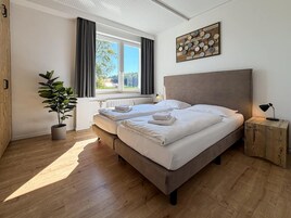 A double bed with white linens and towels, a potted plant, a radiator, and a wall-mounted artwork.
