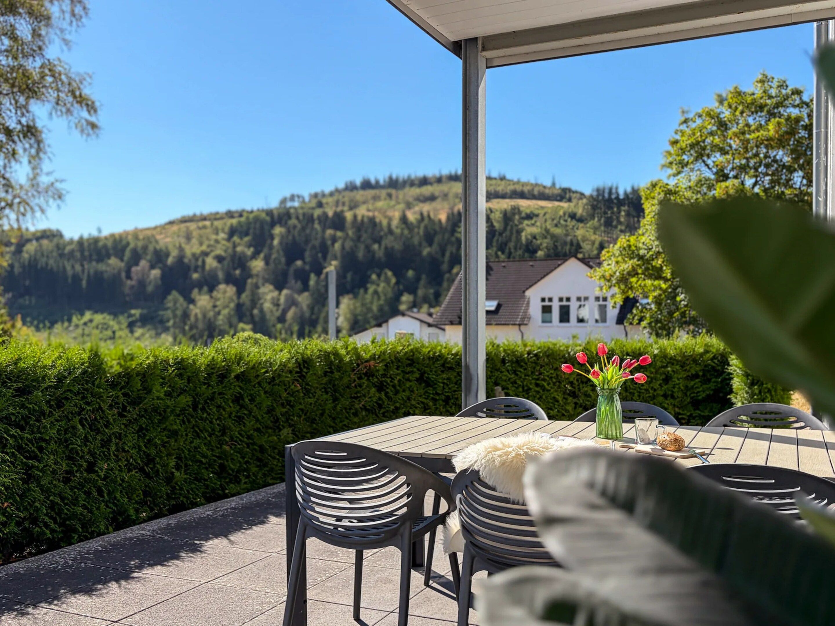 Une terrasse extérieure avec une table et des chaises, un vase rempli de fleurs et une vue sur une maison et une colline boisée.