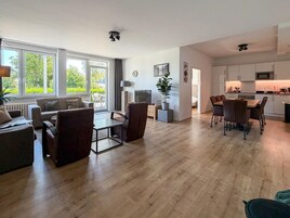 Una sala moderna con sofá, sillones y una mesa de centro. Hay un comedor con una mesa y sillas. La habitación tiene ventanas grandes y vistas a zonas verdes.