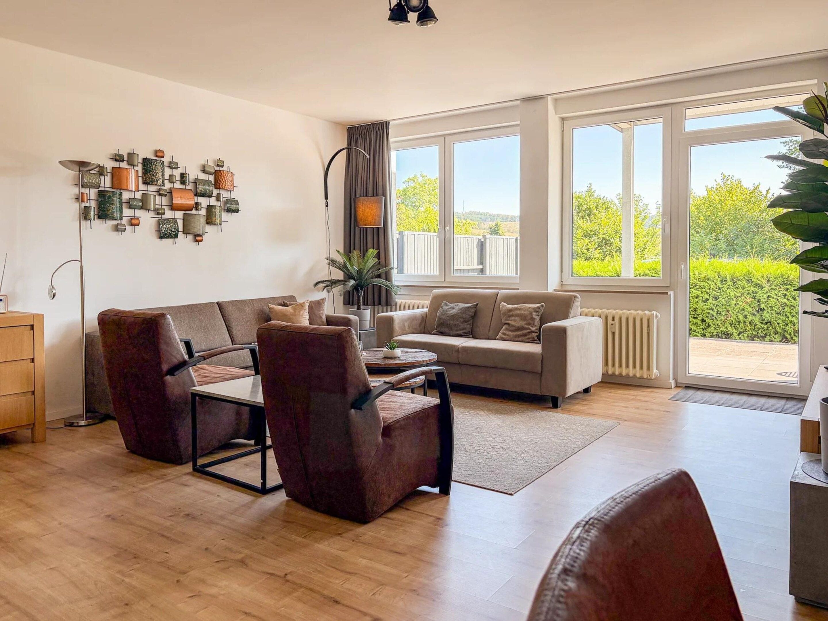 A spacious living room with a sofa, armchairs, and a coffee table. There is a large window providing a view of greenery outside.