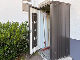Una puerta metálica de seguridad con manija y cerradura, un trapeador apoyado en ella, y un arbusto verde al lado de la puerta.