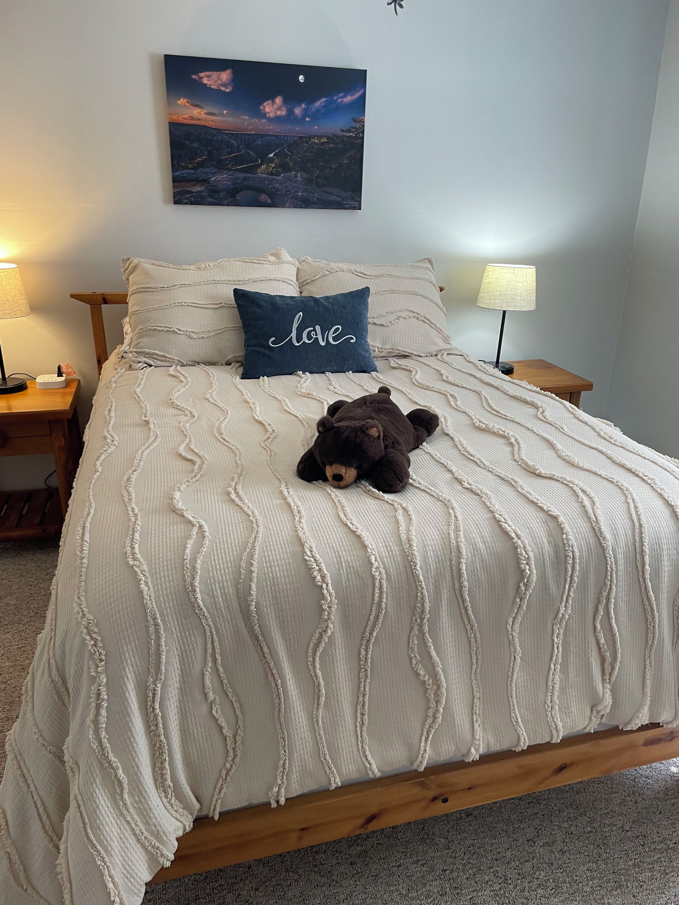 A bedroom with a bed, a teddy bear, a nightstand, a lamp, and a wall-mounted painting.
