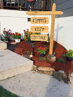 Un segnale di legno con la scritta 'KIMMY'S KOZY COTTAGE' e un'area giardino con piante in vaso e pacciamatura.
