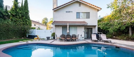 A residential property with a swimming pool, a patio with outdoor furniture, and a two-story house with a tiled roof.