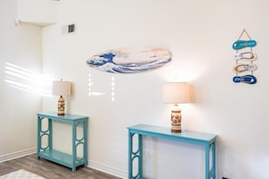 A room with two teal side tables, each with a lamp, a wall-mounted artwork of a wave, and a wall decoration with the phrase 'The Beach Is My Happy Place.'