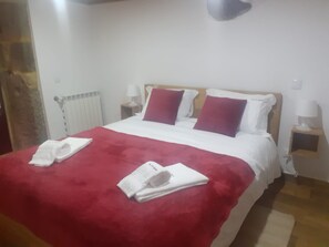 Una cama bien hecha con una manta roja y almohadas, una mesita de noche blanca con una lámpara, y un radiador en la pared.