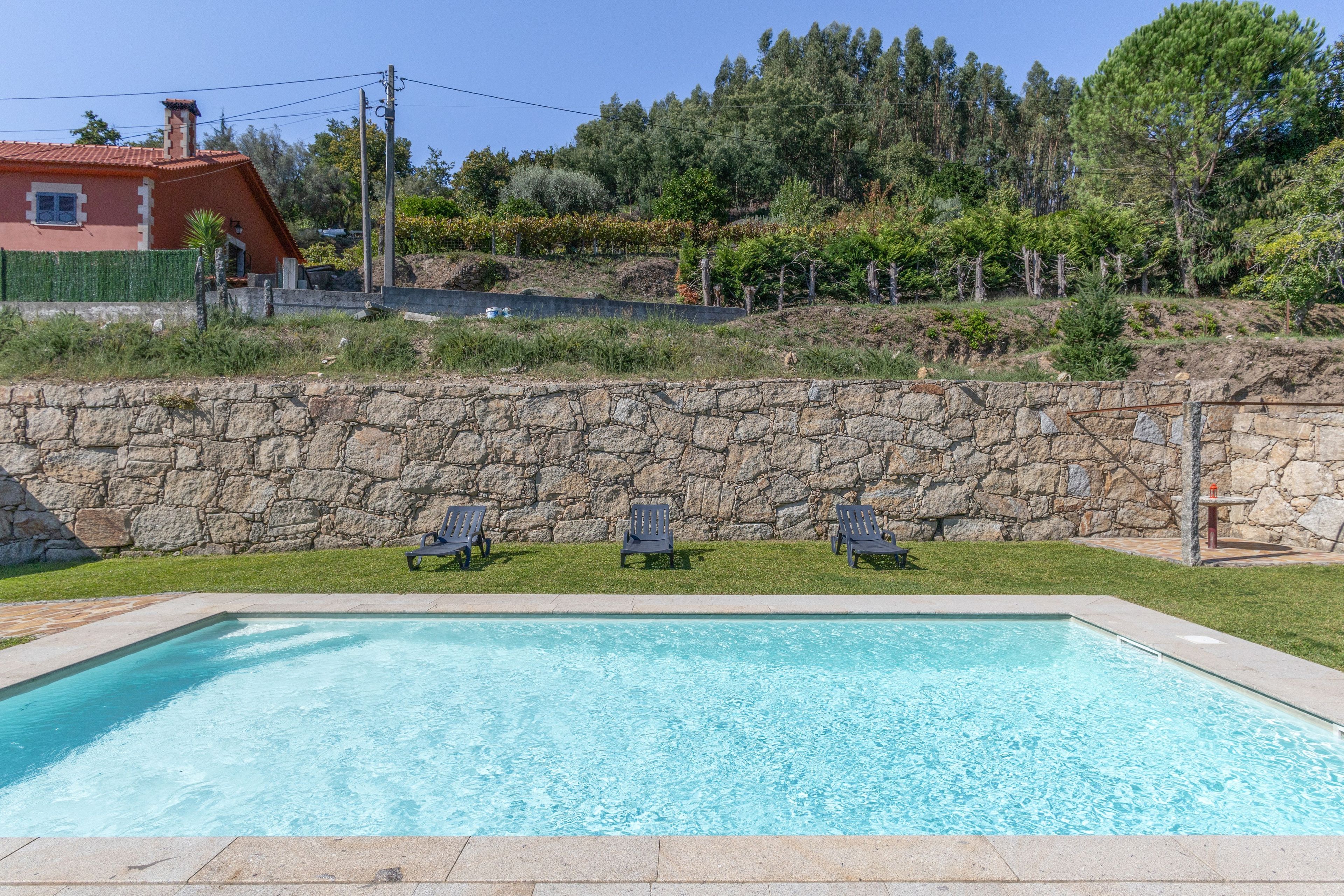 A swimming pool with three lounge chairs and a stone wall in the background.