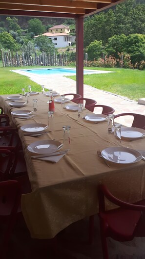 Un área de comedor al aire libre con mesas dispuestas para una comida, con vistas a una piscina y un jardín ajardinado.