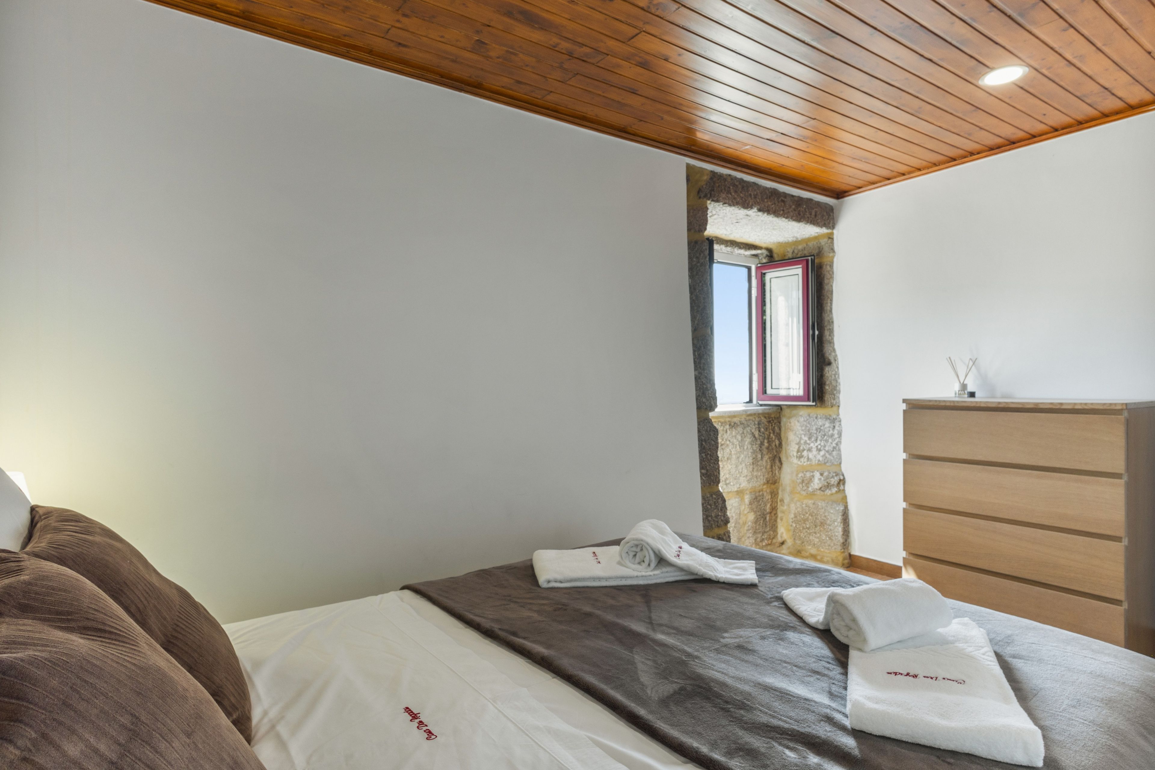 Une chambre avec un plafond en bois, un mur en pierre, une fenêtre, un lit recouvert d’une courtepointe blanche et marron, et une commode en bois.