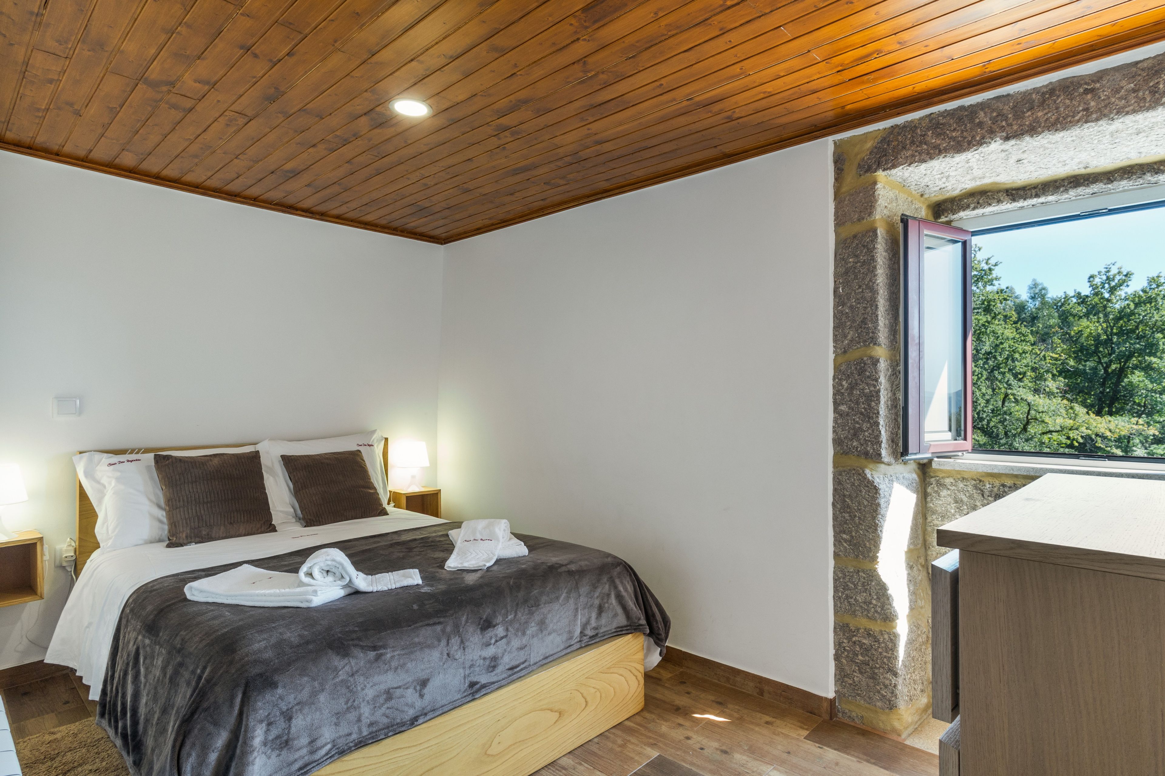 A bedroom with a wooden ceiling, a stone wall, a bed with a grey blanket, and a window with a view of trees.