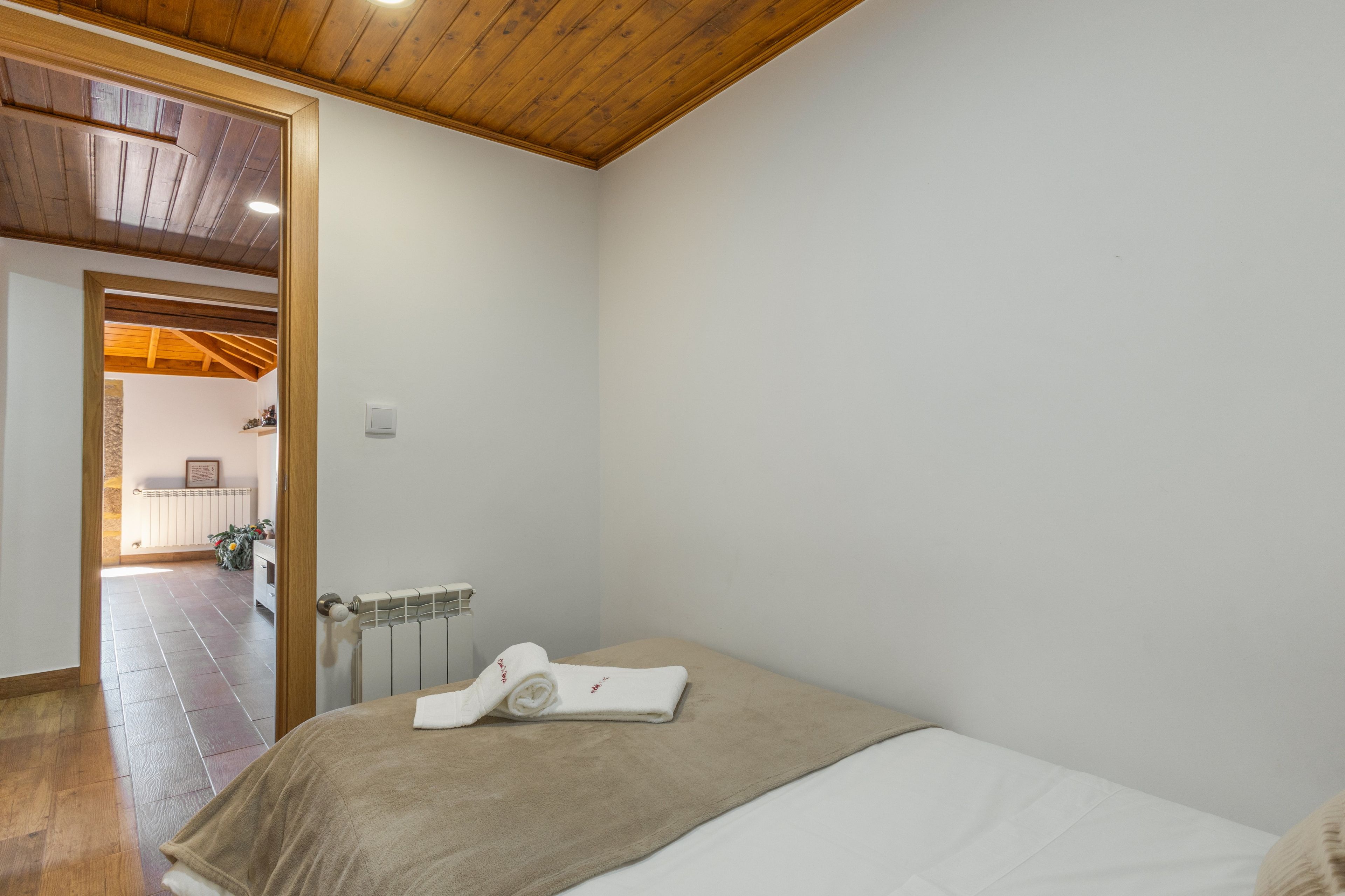 Une chambre avec un plafond en bois, un radiateur, un lit recouvert d’un drap blanc et une serviette.
