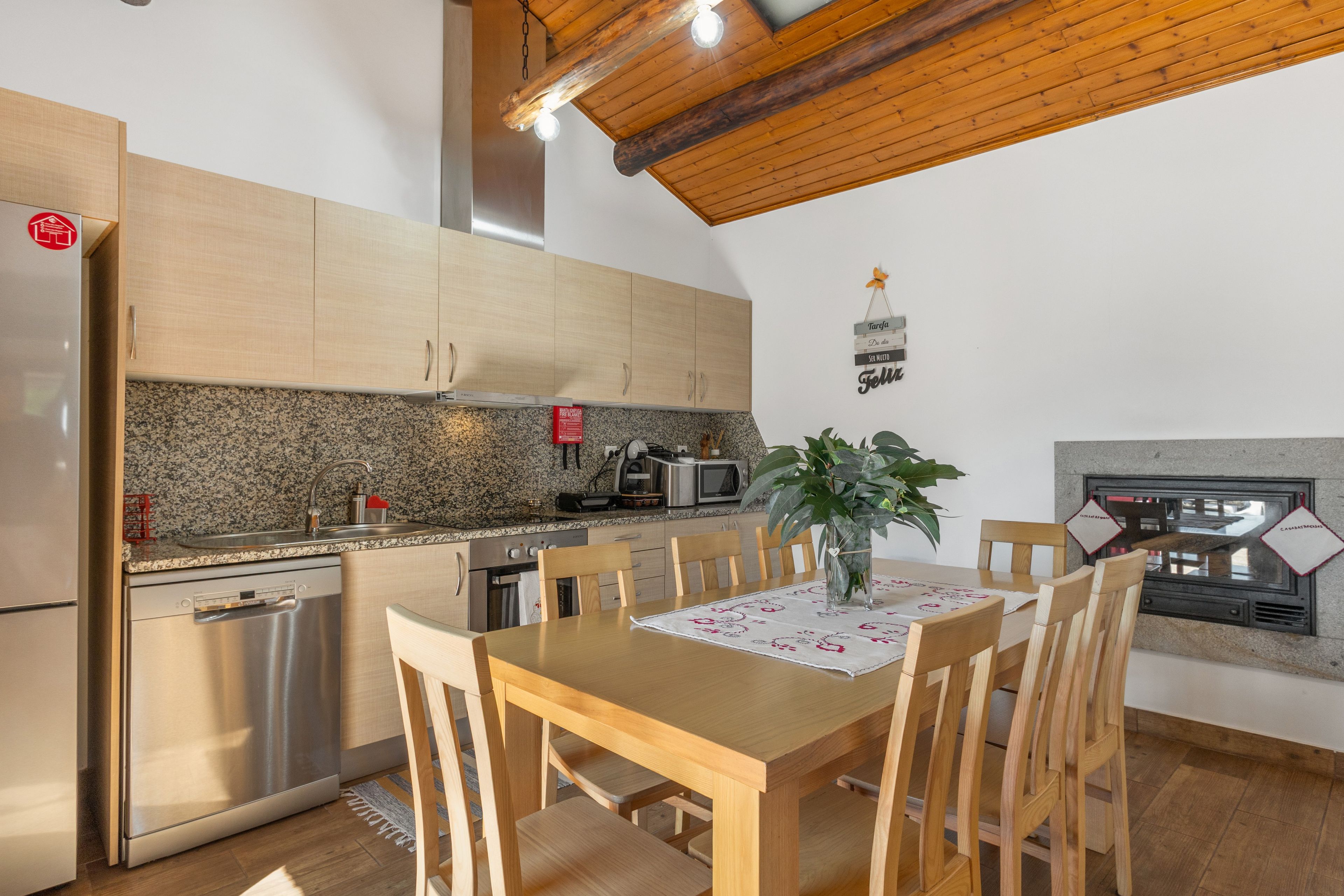 Una cocina con armarios de madera, encimera de granito, lavavajillas de acero inoxidable, una mesa de comedor de madera con sillas, una planta en maceta y un letrero colgante.