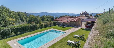 Una piscina con sillas de descanso, una casa de piedra con techo rojo y vistas a las montañas.