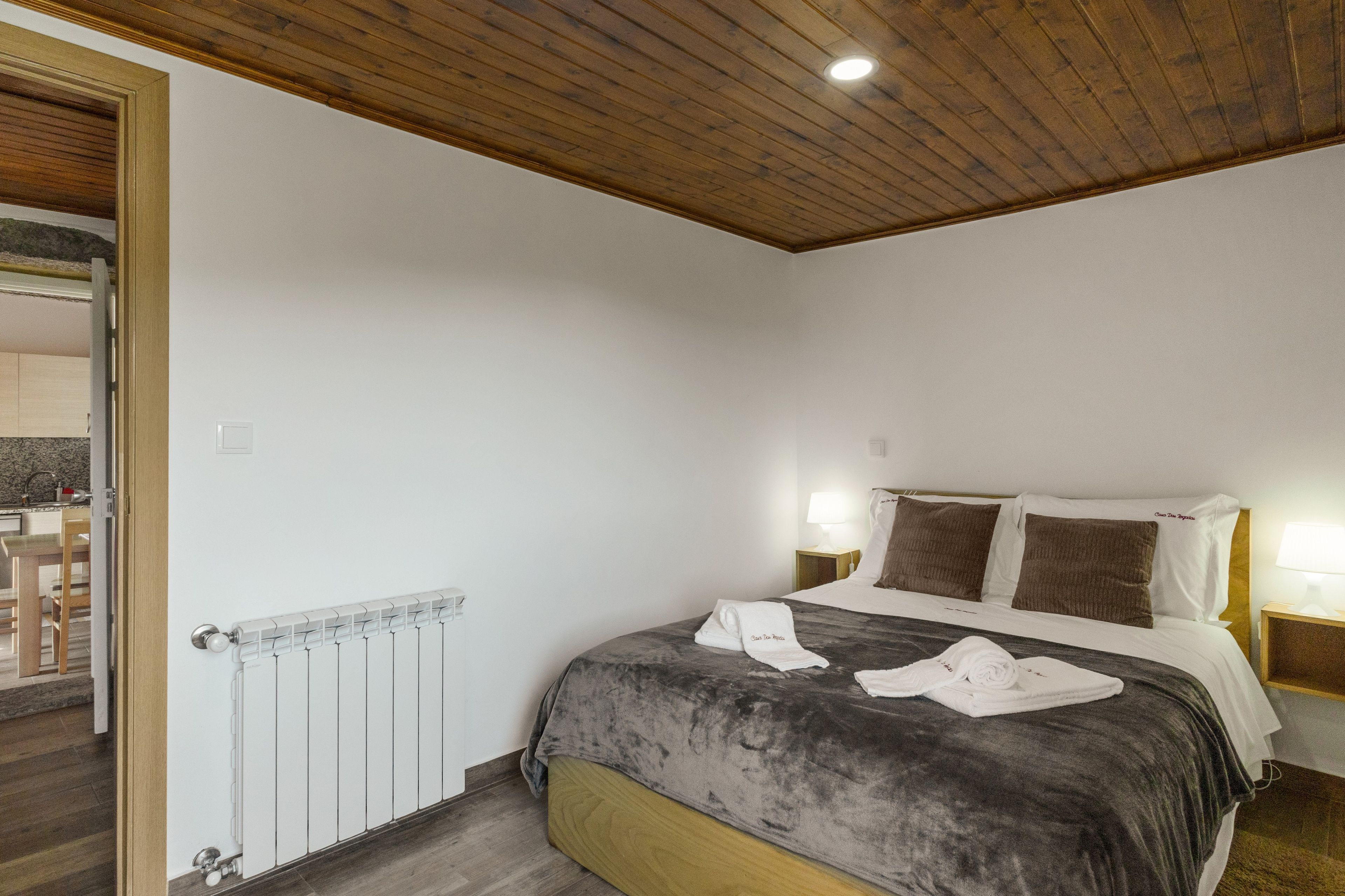A bedroom with a wooden ceiling, a bed with a grey duvet, two bedside lamps, and a radiator.