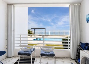 A balcony with a view of a swimming pool and beach, two chairs with patterned cushions, and a fan on the floor. - Ocean Views. Golden Evenings. Unforgettable Stays. (Rincón)